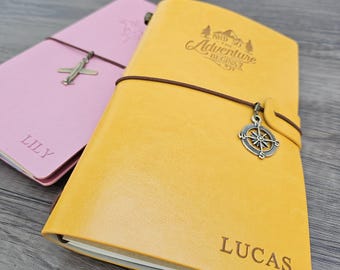 May include: Two leather journals, one pink and one yellow, with brown elastic closures. The pink journal has an airplane charm and the name "LILY" engraved. The yellow journal features a compass charm and the phrase "AND SO THE ADVENTURE BEGINS" with the name "LUCAS" engraved.