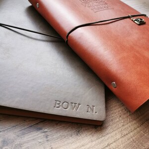 May include: Two leather journals, one brown and one gray, are displayed on a wooden surface. The brown journal has a black cord closure with a charm. The gray journal is embossed with the text "BOW N."