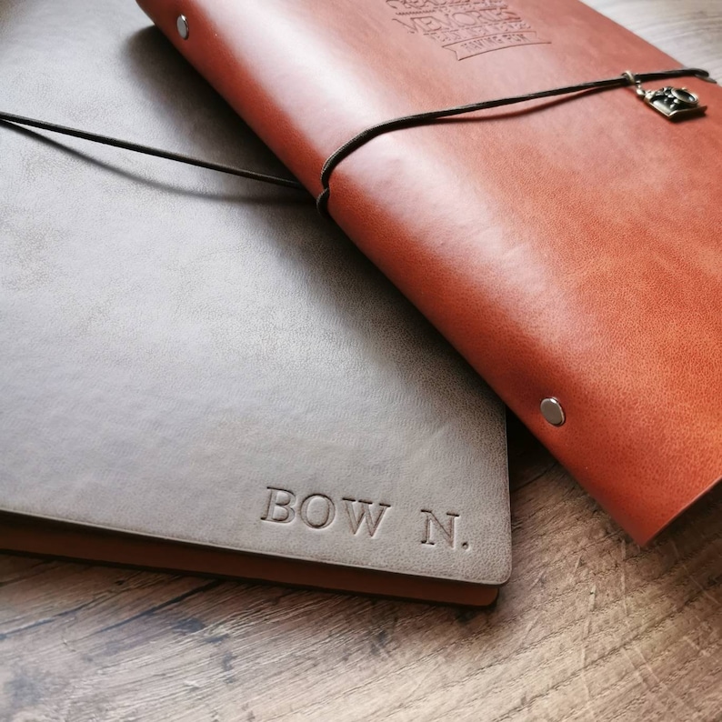 May include: Two leather journals, one brown and one gray, are displayed on a wooden surface. The brown journal has a black cord closure with a charm. The gray journal is embossed with the text "BOW N."