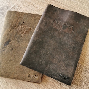 Op de afbeelding: Twee bruine lederen dagboeken met gepersonaliseerde namen, "ADAM" en "JOSH", gegraveerd op de omslagen. Een dagboek heeft een kompasontwerp, terwijl het andere een natuurscène met bomen en dieren heeft.