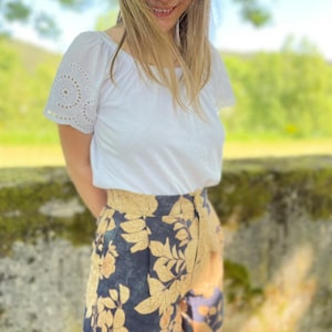 May include: A person wearing a white short-sleeved shirt with eyelet detailing on the sleeves and dark blue shorts with a gold floral print. The shorts have a high waist and a relaxed fit. The person is standing in front of a green mossy wall. The text '@melinutelli' is visible in the lower right corner of the image.