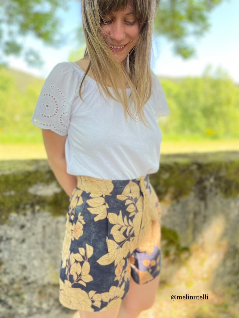 May include: A person wearing a white short-sleeved shirt with eyelet detailing on the sleeves and dark blue shorts with a gold floral print. The shorts have a high waist and a relaxed fit. The person is standing in front of a green mossy wall. The text '@melinutelli' is visible in the lower right corner of the image.