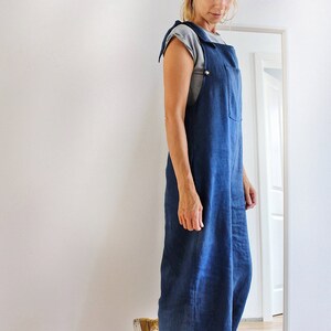Op de afbeelding: Een vrouw draagt een blauwe denim overalljurk met een grijs t-shirt eronder. Ze draagt gele en witte sneakers.