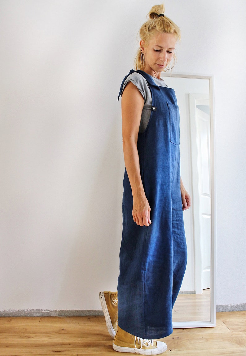 Op de afbeelding: Een vrouw draagt een blauwe denim overalljurk met een grijs t-shirt eronder. Ze draagt gele en witte sneakers.