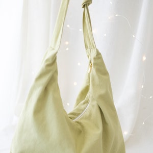 Pode incluir: Uma bolsa tote de lona verde claro com fecho de correr e uma al&ccedil;a de n&oacute;. A bolsa est&aacute; sobre uma superf&iacute;cie branca com &oacute;culos de sol e um grampo de cabelo.