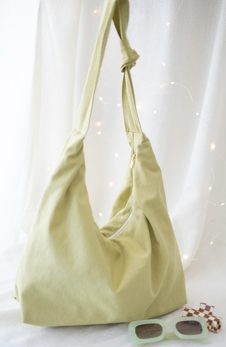 Pode incluir: Uma bolsa tote de lona verde claro com fecho de correr e uma al&ccedil;a de n&oacute;. A bolsa est&aacute; sobre uma superf&iacute;cie branca com &oacute;culos de sol e um grampo de cabelo.