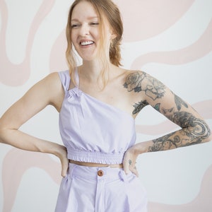 May include: A person wearing a lavender crop top and matching pants. The top has a one-shoulder design and the pants have a high waist. The person has a tattoo on their arm.