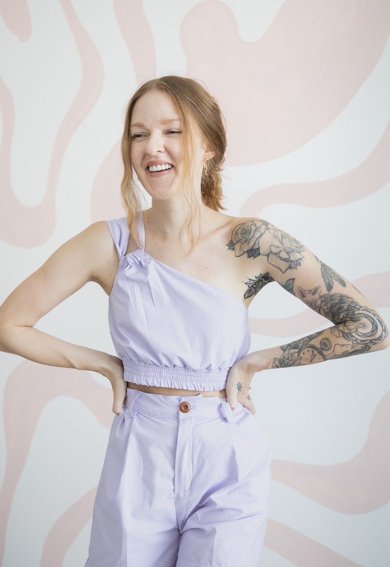 May include: A person wearing a lavender crop top and matching pants. The top has a one-shoulder design and the pants have a high waist. The person has a tattoo on their arm.