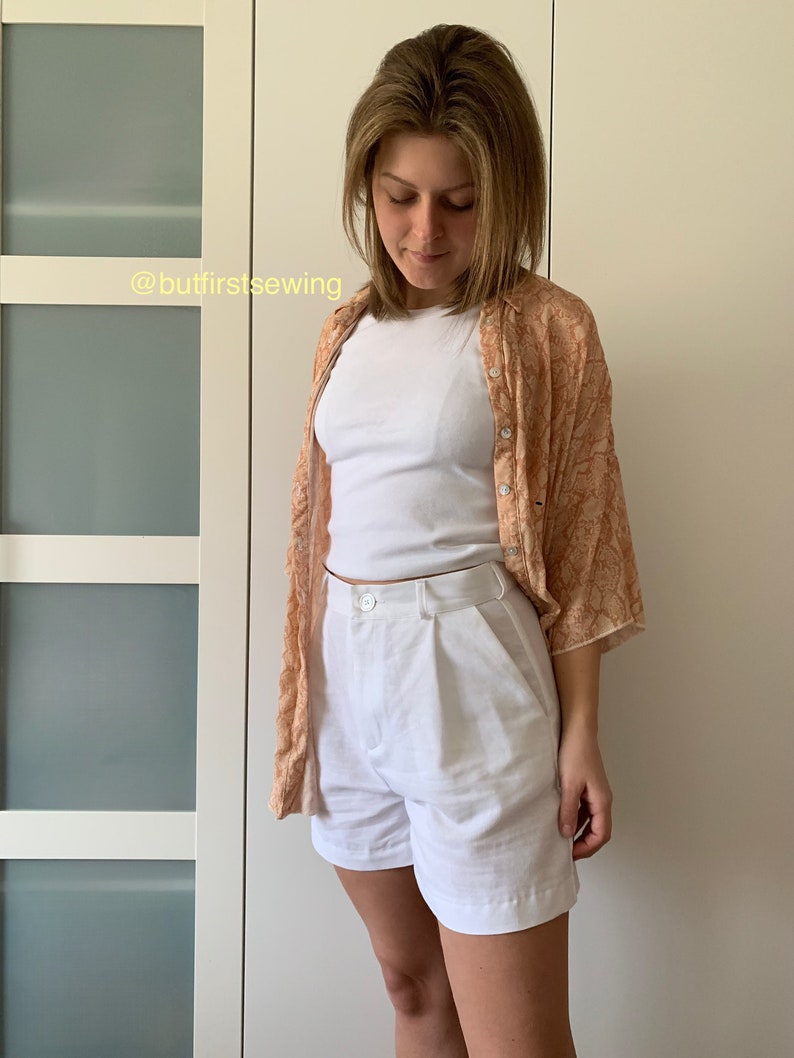 May include: White linen shorts with a button and zipper closure. The shorts have pleats in the front and are worn with a white t-shirt and a peach floral patterned shirt. The text '@butfirstsewing' is visible in the image.