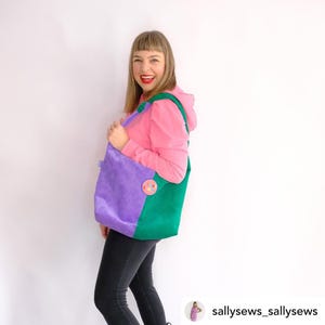 May include: A woman is wearing a pink hoodie and black pants. She is holding a purple and green corduroy tote bag with a colorful patch on the front. The bag has a long strap.