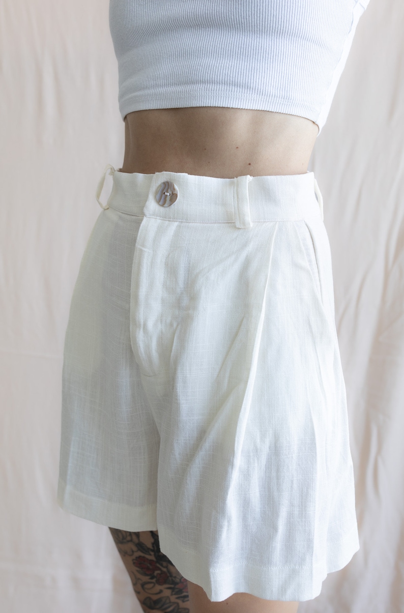 May include: White linen shorts with a button closure and pleats.