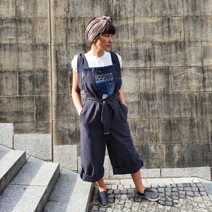 Op de afbeelding: Een vrouw draagt een donkerblauwe denim jumpsuit met een patchwork ontwerp op de borst. Ze draagt er een wit shirt onder en een roze hoofdband. Ze staat voor een grijze bakstenen muur.