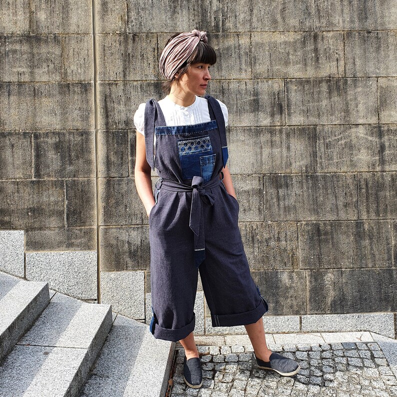 Op de afbeelding: Een vrouw draagt een donkerblauwe denim jumpsuit met een patchwork ontwerp op de borst. Ze draagt er een wit shirt onder en een roze hoofdband. Ze staat voor een grijze bakstenen muur.