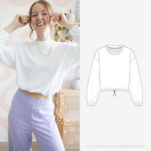 Può includere: Felpa bianca corta con maniche lunghe e coulisse sul fondo. La felpa è mostrata su una modella che la indossa con pantaloni color lilla chiaro.