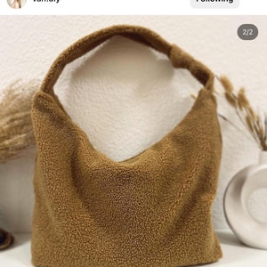Pode incluir: Uma bolsa de ombro marrom e felpuda feita de tecido sherpa. A bolsa tem uma al&ccedil;a &uacute;nica e uma forma curva.