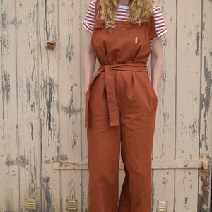 Op de afbeelding: Een roestkleurige jumpsuit met een brede, bruine riem en een gestreept wit en rood T-shirt eronder. De jumpsuit heeft grote zakken aan de voorkant en een ontspannen pasvorm. De persoon draagt gele sneakers.