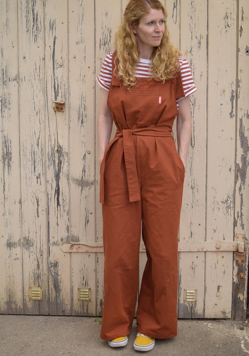 Op de afbeelding: Een roestkleurige jumpsuit met een brede, bruine riem en een gestreept wit en rood T-shirt eronder. De jumpsuit heeft grote zakken aan de voorkant en een ontspannen pasvorm. De persoon draagt gele sneakers.