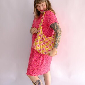 May include: A woman wearing a pink floral dress with short sleeves is holding a yellow and pink floral fabric tote bag with a long strap. The bag is slung over her shoulder. The woman has tattoos on her arms and legs.