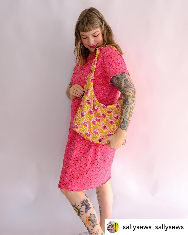 May include: A woman wearing a pink floral dress with short sleeves is holding a yellow and pink floral fabric tote bag with a long strap. The bag is slung over her shoulder. The woman has tattoos on her arms and legs.