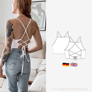 Puede incluir: Top blanco corto con un detalle de encaje cruzado en la espalda. El top se muestra en una modelo que lleva jeans azules. La imagen también incluye un dibujo de línea del top.