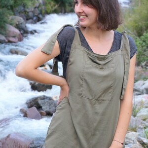 Op de afbeelding: Een vrouw draagt een groene overall met een gestreept shirt eronder. Ze staat voor een rotsachtige beek.