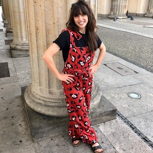 Op de afbeelding: Een vrouw draagt een rood en zwart luipaardprint jumpsuit met een zwart t-shirt eronder. Ze staat voor een stenen pilaar.