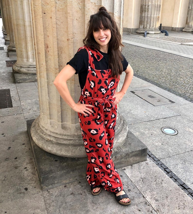 Op de afbeelding: Een vrouw draagt een rood en zwart luipaardprint jumpsuit met een zwart t-shirt eronder. Ze staat voor een stenen pilaar.