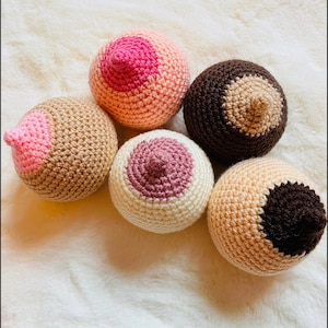 Op de afbeelding: Een verzameling van vijf gehaakte borstvormige objecten in verschillende huidtinten. Elk heeft een tepel en tepelhof in tinten roze, bruin en beige. De objecten zijn gerangschikt op een zacht, wit, getextureerd oppervlak.