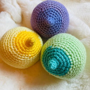 Puede incluir: Tres esferas de ganchillo en tonos amarillo, morado y verde, cada una con una protuberancia similar a un pezón. Las esferas están dispuestas sobre una superficie suave y blanca texturizada. El trabajo de ganchillo es detallado y los colores son vibrantes.