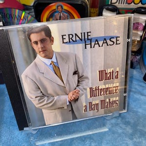 May include: CD case featuring Ernie Haase in a light gray suit and gold tie. The album title "What a Difference a Day Makes" is displayed in white text. The background includes other CDs and Sesame Street memorabilia.