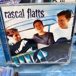 May include: A CD case with a blue background featuring a band called "Rascal Flatts". The band members are pictured in a car, with the lead singer in the front seat and the other two members in the back.