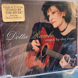 May include: CD case featuring Dottie Rambo with a guitar. The album cover includes the text "STAND BY THE RIVER" featuring Dolly Parton. The album cover has a brown and red color scheme.