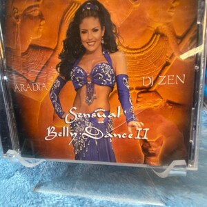 May include: A CD case with a woman in a blue and silver belly dancing costume. The background is a gold and orange design with the text "Aradía" and "DJ Zen". The text "Sensual Belly Dance II" is also on the case.