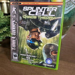 May include: A green Xbox game case for "Splinter Cell: Chaos Theory" featuring Tom Clancy's name. The cover art shows a man in tactical gear. The case has the Xbox logo and the Ubisoft logo.