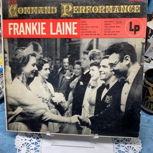May include: A vintage Frankie Laine "Command Performance" LP record album cover. The album cover has a red banner with the artist's name, "FRANKIE LAINE", in white. The album cover features a black and white photo of people in formal attire.