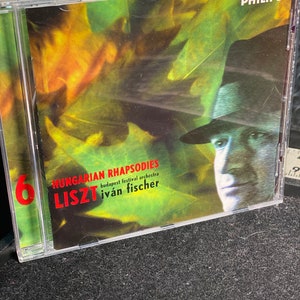 May include: A CD case with a black and white photo of a man wearing a hat. The text on the case reads "Hungarian Rhapsodies Liszt Budapest Festival Orchestra Ivan Fischer".