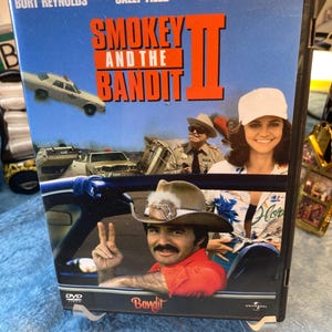 May include: DVD case for the movie "Smokey and the Bandit II" featuring Burt Reynolds, Sally Field, and Jackie Gleason. The cover has a blue background with images of cars and the actors. The title is in large orange letters.