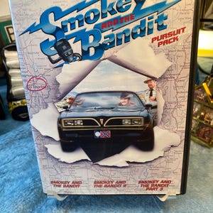 May include: DVD box set for the movie "Smokey and the Bandit: Pursuit Pack." The cover features a black car with a Confederate flag, a map background, and the movie title in blue. The set includes parts 1, 2, and 3.
