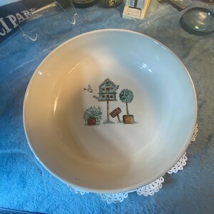 May include: A white ceramic bowl with a blue rim features a whimsical design of a birdhouse with birds, trees, and signs. The bowl is on a white lace doily.