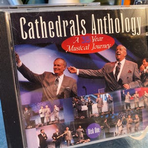 May include: A CD case featuring the album "Cathedrals Anthology: A 35 Year Musical Journey." The cover shows two men in suits with microphones, and a collage of other images. The words "Disk One" are visible.