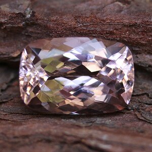 May include: A faceted, pink gemstone with a rectangular shape. The gemstone has a smooth, polished surface and is set against a brown, textured background.