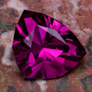 Könnte beinhalten: Ein facettierter, dreieckiger Edelstein in leuchtendem Magenta. Der Schliff des Steins zeigt mehrere Innenfacetten, die ein Spiel von Licht und Schatten erzeugen. Der Edelstein liegt auf einer marmorierten, rotbraunen Oberfläche.