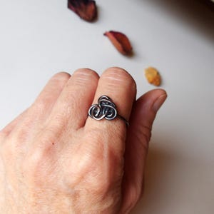 May include: A silver-toned ring with a looped design is worn on a finger. The ring's focal point is a cluster of intertwined metal loops. The background includes dried leaves on a white surface, creating a minimalist aesthetic.