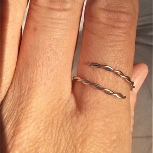 May include: A silver-colored, adjustable ring with a twisted design. The ring wraps around the finger with two parallel, textured bands. The ring is shown on a hand, highlighting the jewelry's design and fit.