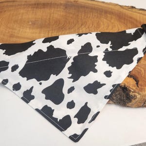 Peut inclure: Un bandana triangulaire pour animal de compagnie avec un motif de taches de vache noir et blanc. Le tissu est fixé à un collier noir réglable doté d'une boucle en plastique à ouverture latérale et d'un anneau en D en métal pour la laisse.