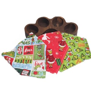Op de afbeelding: Een houten serveerblad met vier cirkelvormige uitsparingen, met drie kerstservetten van stof. De servetten hebben Grinch-personages en feestelijke patronen in rood, groen en wit. De servetten zijn in een driehoek gevouwen.