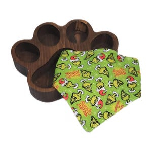 Könnte beinhalten: Ein dunkelbraunes, pfotenförmiges Holzserviertablett mit fünf kreisförmigen Aussparungen. Ein grünes Hunde-Bandana mit einem sich wiederholenden Muster des Grinch mit Weihnachtsmütze und dem Text "Feelin' Grinchy" liegt neben dem Tablett.