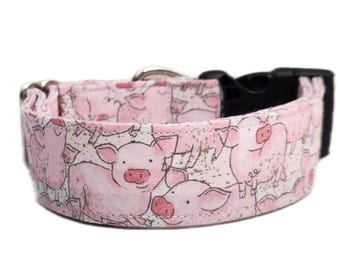 May include: A pink fabric pet collar featuring a repeating pattern of illustrated pigs with dark outlines. The collar includes a black plastic side-release buckle, a silver metal D-ring for leash attachment, and a silver metal adjustable slider.