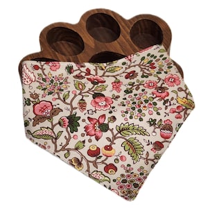 Pode incluir: Um suporte de madeira para ovos com seis reentrâncias circulares, combinado com uma bandana com padrão floral. A bandana apresenta um fundo bege com um padrão repetido de flores e folhas rosa, vermelhas e verdes.