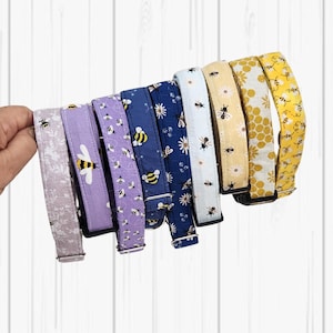 May include: A selection of eight dog collars, each patterned with bees and flowers. The collars are in a variety of colours, including purple, blue, and yellow. Each collar has a metal buckle and is held in a hand.